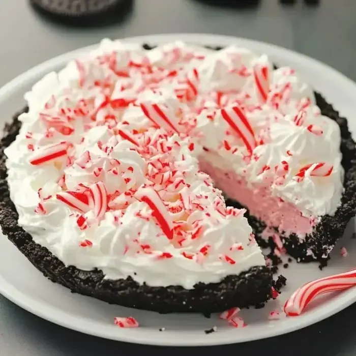 A festive peppermint pie with a chocolate crust, topped with whipped cream and crushed candy canes, with a slice removed.