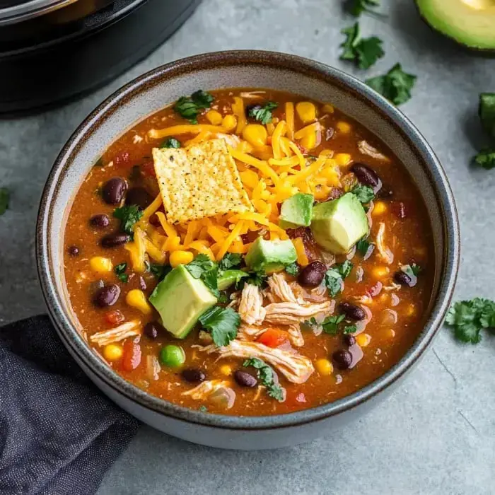 A bowl of hearty soup featuring shredded chicken, black beans, corn, diced avocado, cheddar cheese, and tortilla chips, garnished with cilantro.