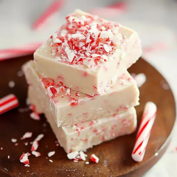 A stack of creamy white peppermint fudge topped with crushed candy canes, beside small candy canes on a wooden platter.