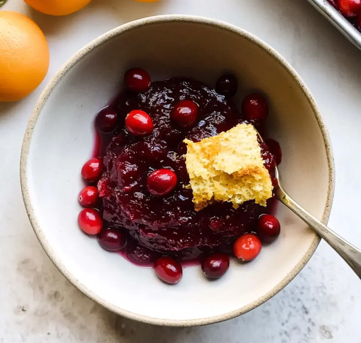 A bowl of cranberry sauce topped with fresh cranberries and a piece of cornbread, accompanied by whole oranges in the background.