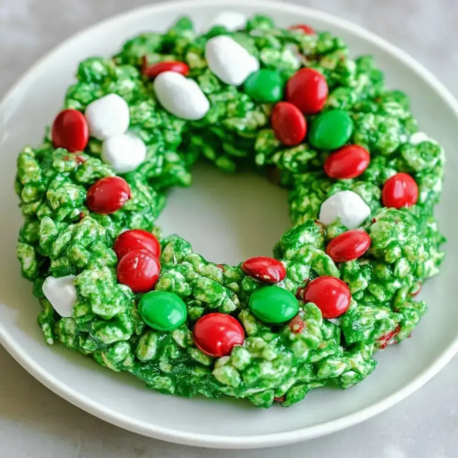 A festive green popcorn wreath decorated with red and green candies and white marshmallows on a plate.