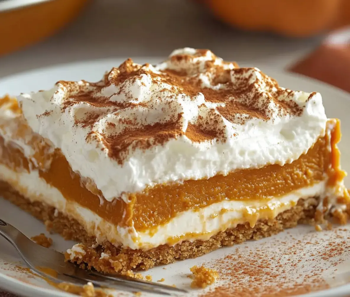 A slice of pumpkin dessert layered with whipped cream and a graham cracker crust, garnished with a sprinkle of cinnamon.