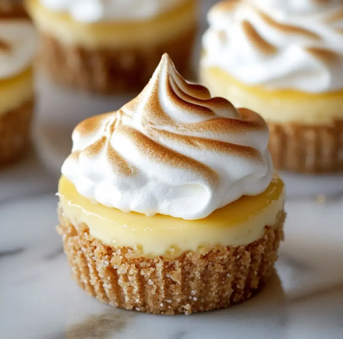 A close-up of a lemon meringue cupcake with a golden brown toasted meringue topping and a graham cracker crust.