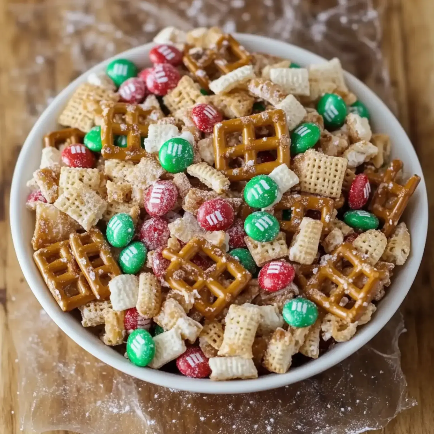 A close-up of a bowl filled with a colorful mix of snack cereals, pretzels, and red and green chocolate candies.