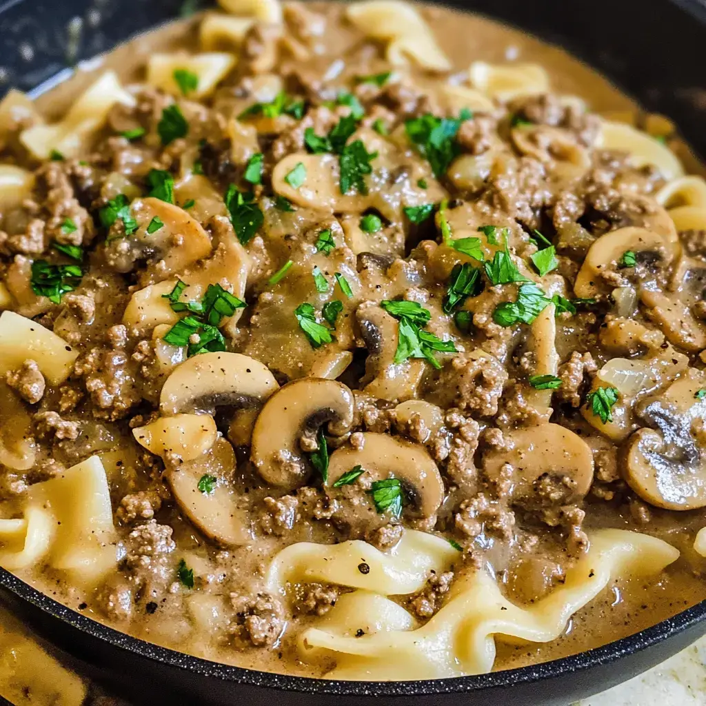 A skillet filled with egg noodles topped with a creamy mixture of ground beef, mushrooms, and chopped parsley.