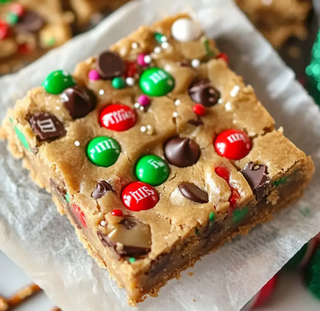 A close-up of a festive dessert bar topped with colorful M&M's and chocolate chips on a piece of parchment paper.