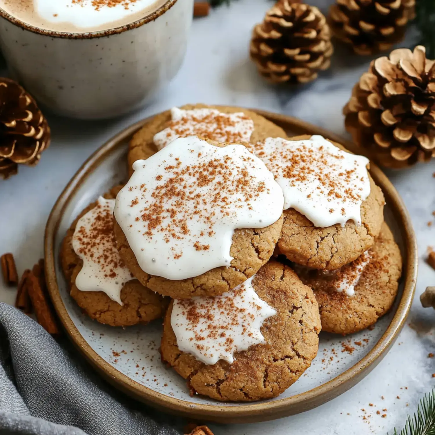 A plate of cookies topped with white frosting and sprinkled with cinnamon, accompanied by a cup of hot chocolate and pine cones.