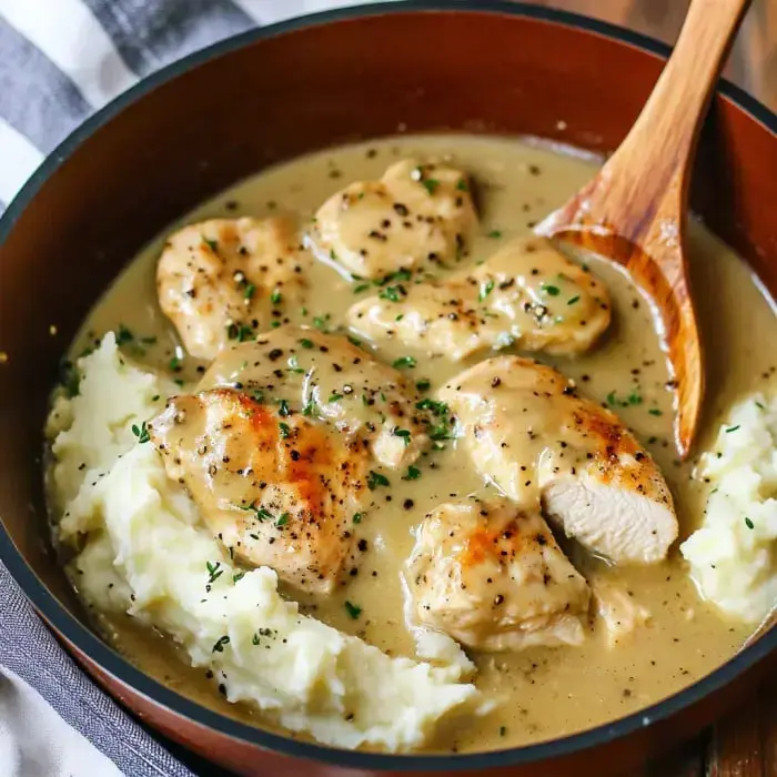 A bowl of creamy chicken gravy with tender pieces of chicken served over a bed of mashed potatoes, garnished with herbs.