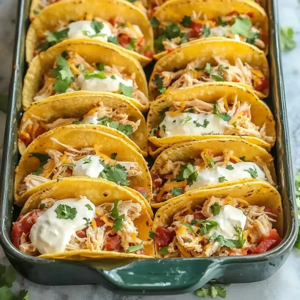 A tray of filled tacos topped with shredded chicken, cheese, diced tomatoes, sour cream, and fresh cilantro.