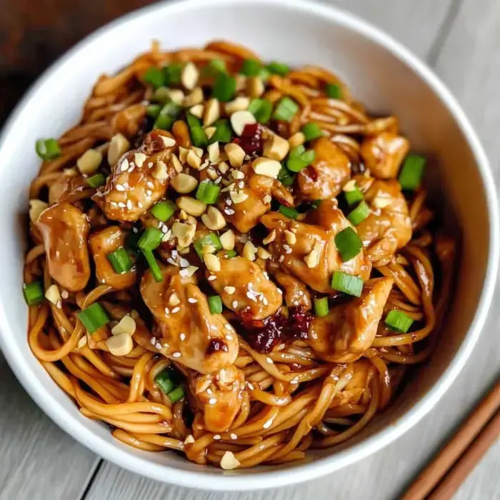 A bowl of chicken and noodles topped with chopped peanuts, green onions, and sesame seeds.