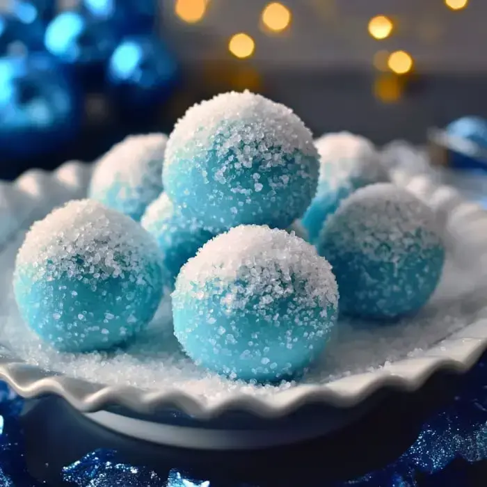 A plate of blue sugar-coated spheres dusted with granulated sugar is set against a festive backdrop of blue ornaments and soft lighting.