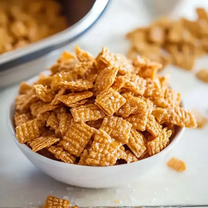 A bowl filled with crunchy, golden-brown cereal pieces sits on a white surface, surrounded by scattered pieces.