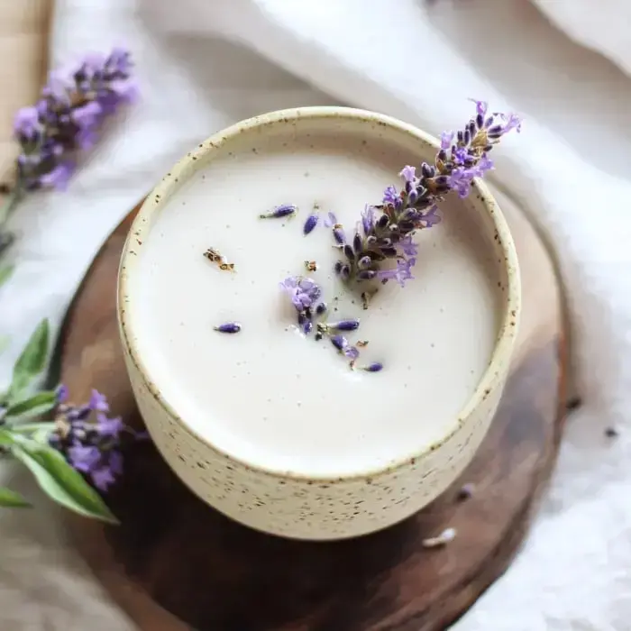 A top-down view of a creamy beverage garnished with fresh lavender flowers in a speckled ceramic cup, resting on a wooden surface.