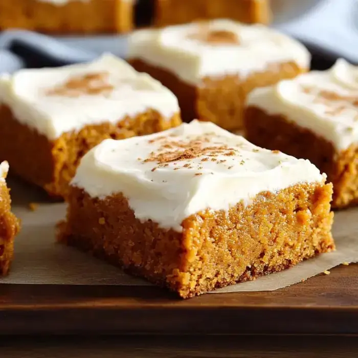 A close-up of pumpkin bars topped with cream cheese frosting and a sprinkle of cinnamon, arranged on a wooden board.