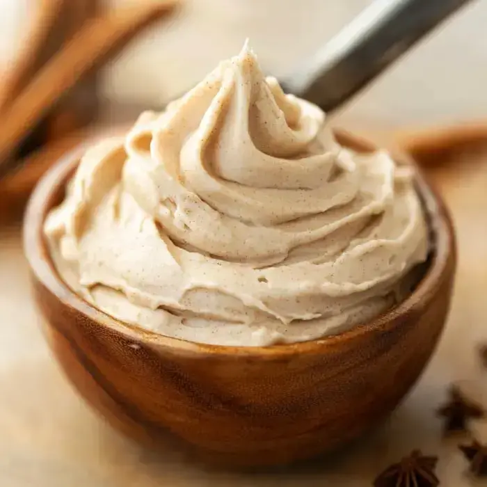 A creamy, light brown buttercream frosting is elegantly swirled in a wooden bowl, surrounded by cinnamon sticks.