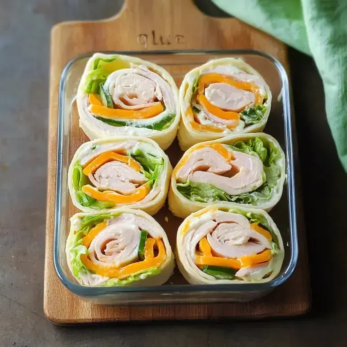 A glass dish holds six roll-ups made with tortillas, turkey, lettuce, and colorful bell peppers arranged neatly.