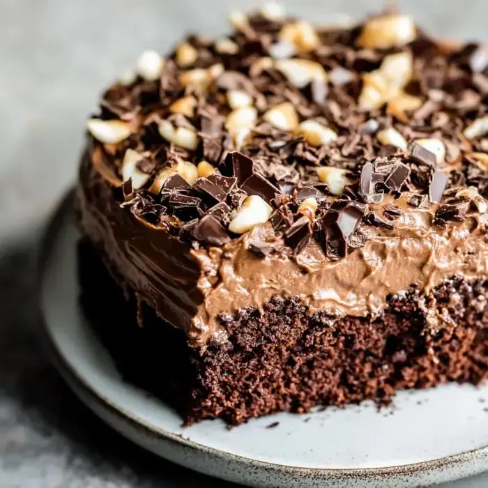 A slice of rich chocolate cake topped with chocolate shavings and nuts, served on a plate.