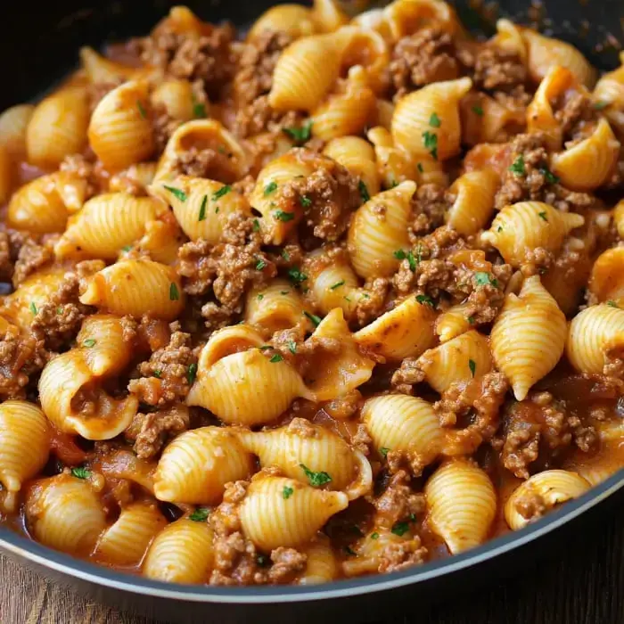 A pan filled with cooked shell pasta mixed with ground beef and a rich tomato sauce, garnished with parsley.