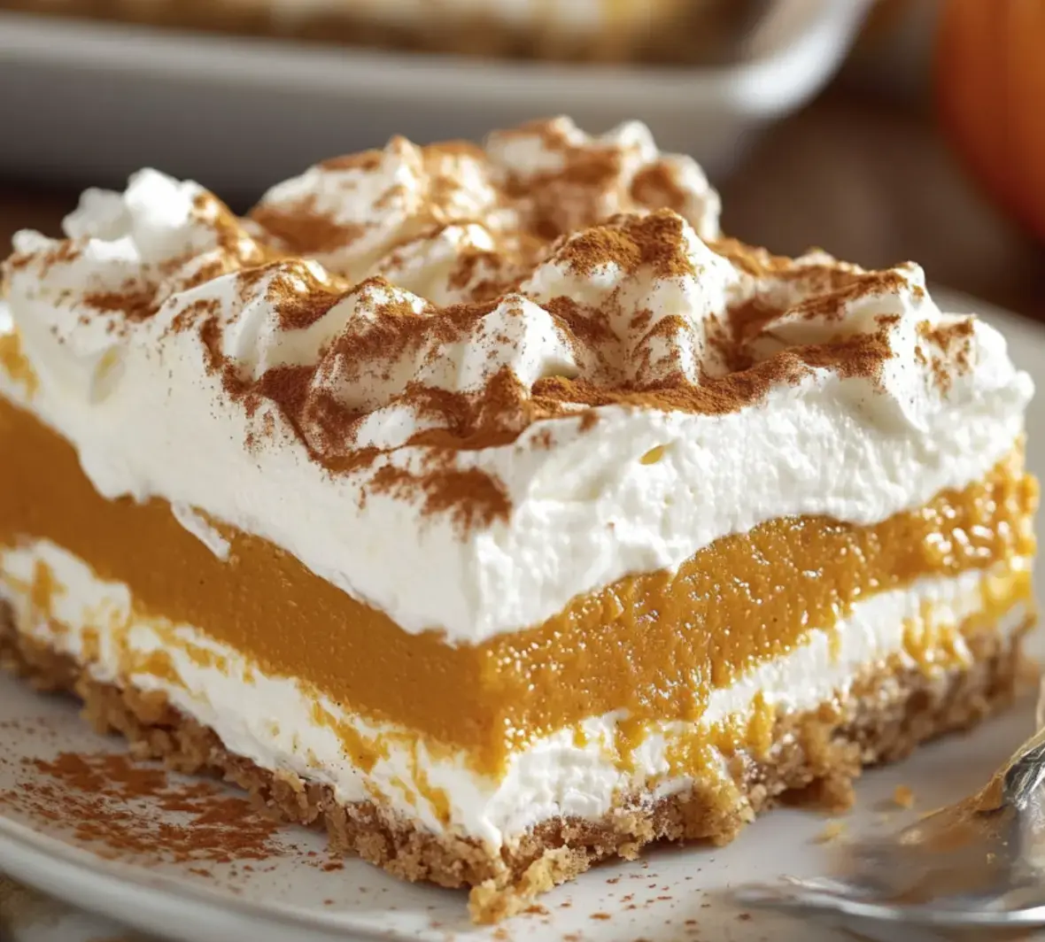 A close-up of a slice of pumpkin dessert layered with whipped cream and a graham cracker crust, topped with a dusting of cinnamon.