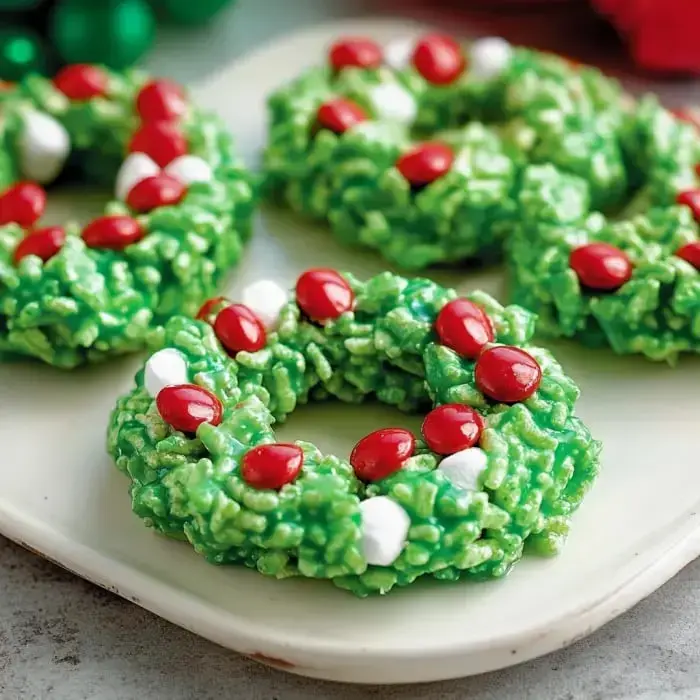 A plate of festive green rice cereal treats shaped like wreaths, decorated with red candies and white marshmallows.