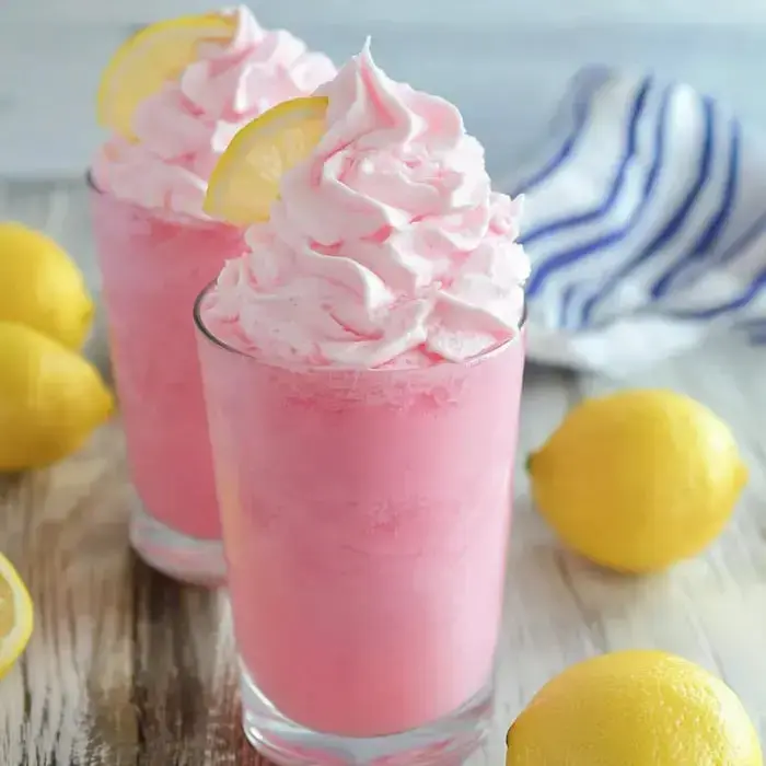 Two pink frosted drinks topped with whipped cream and lemon slices are displayed alongside fresh lemons on a wooden surface.