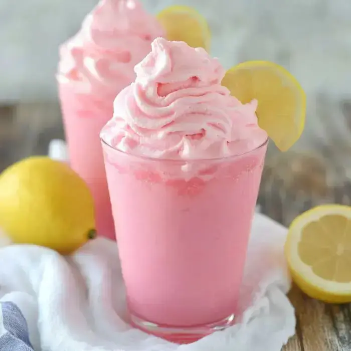 Two glasses of pink whipped lemon drink are garnished with lemon slices, accompanied by fresh lemons on a light cloth.