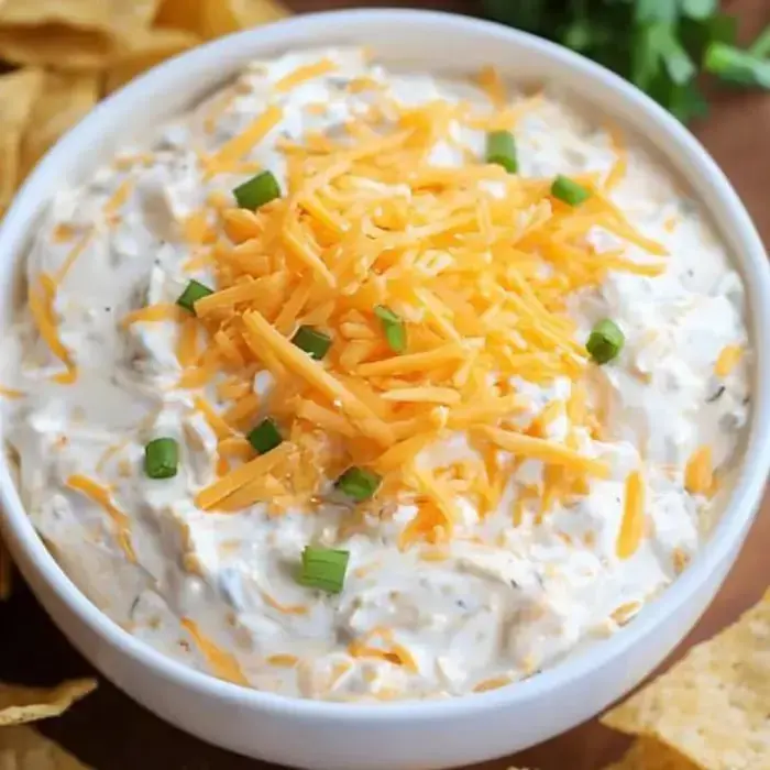 A bowl of creamy dip topped with shredded cheddar cheese and green onions, accompanied by tortilla chips.