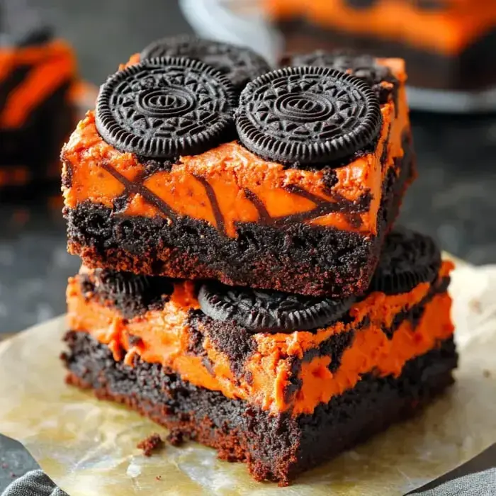 Two stacked brownie squares topped with orange frosting and Oreo cookies, set against a dark background.