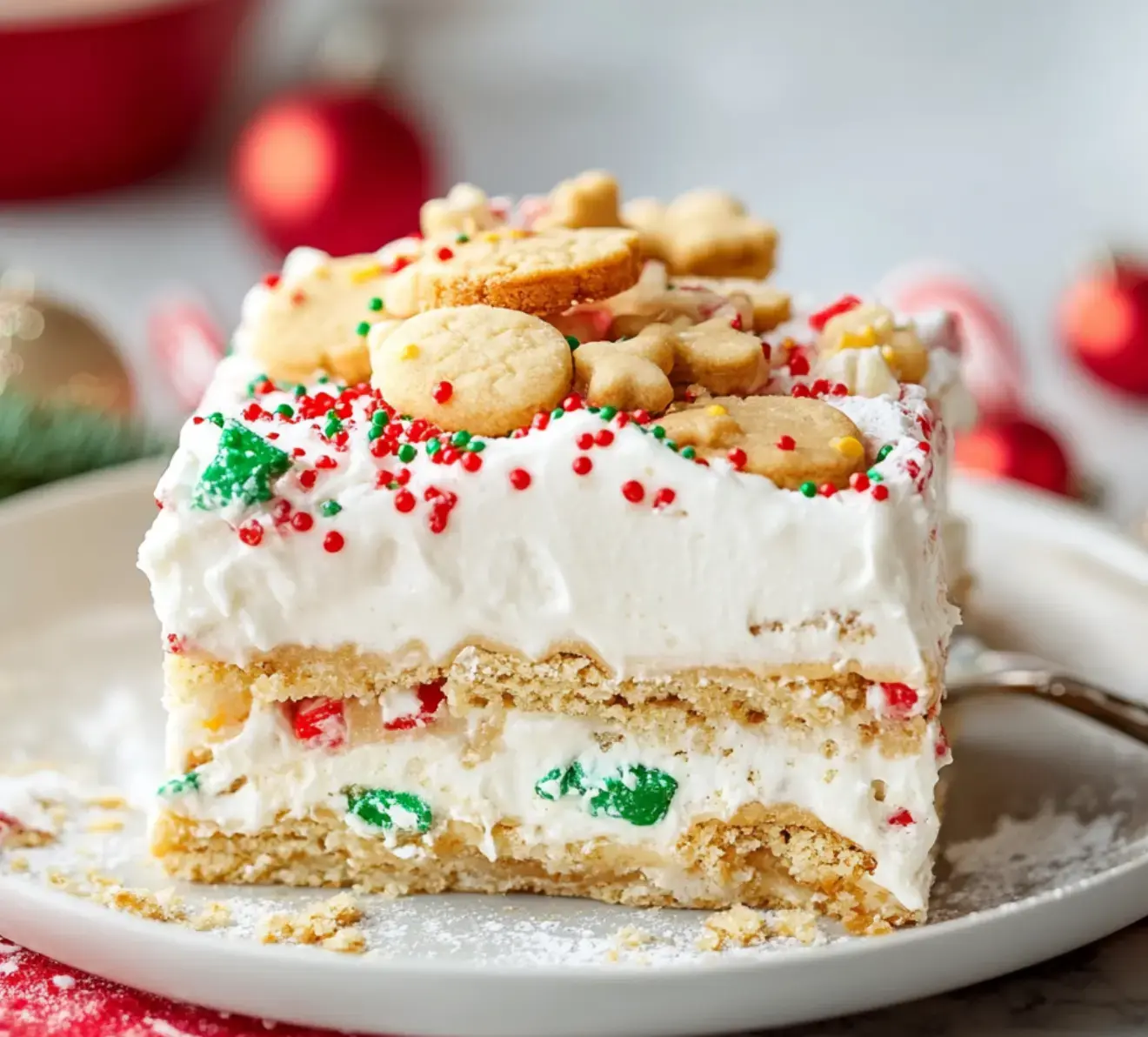 A decorated holiday cake with layers of cream, colorful sprinkles, and festive cookie shapes on top is displayed on a plate.