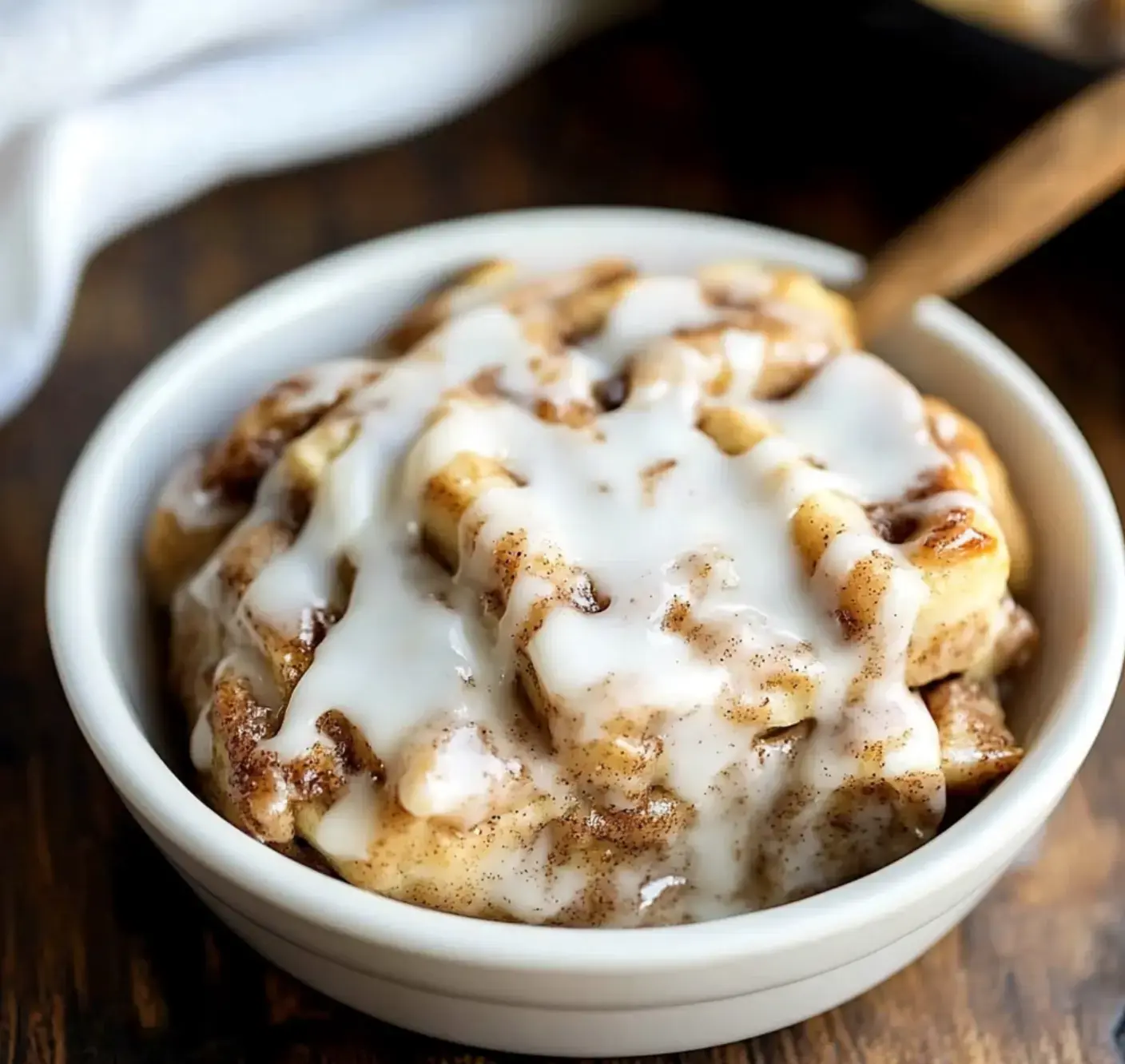 A bowl of cinnamon rolls drizzled with white icing sits on a wooden surface.