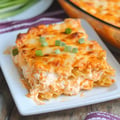 A serving of creamy lasagna topped with green onions on a white plate, with a baking dish of lasagna in the background.