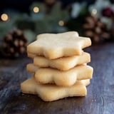 A stack of four star-shaped cookies is arranged on a wooden surface, with blurred pinecones and festive greenery in the background.