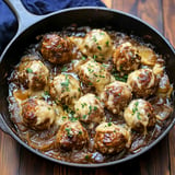 A cast iron skillet filled with meatballs covered in melted cheese and sprinkled with fresh parsley, sitting in a flavorful brown sauce.