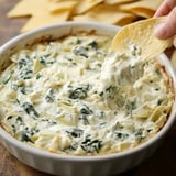 A hand is dipping a tortilla chip into a creamy, cheesy spinach and artichoke dip in a white baking dish, with additional chips in the background.