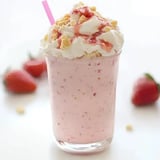 A pink milkshake topped with whipped cream, crushed cookies, and strawberry syrup, served with a straw, alongside fresh strawberries.