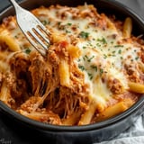 A fork is lifting a portion of cheesy baked pasta with ground meat and tomato sauce from a dark bowl.