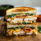 A stacked sandwich filled with grilled chicken, spinach, bacon, and melted cheese, served on toasted bread.