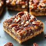 A close-up of a pecan pie bar topped with halved pecans on a dark surface.