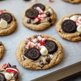 A close-up of freshly baked cookies topped with Oreo pieces, white chocolate chips, and crushed peppermint candies on a baking sheet.