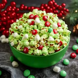 A large green bowl filled with green popcorn mixed with red and white M&M's and small marshmallows, surrounded by red festive decorations.