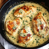 A skillet with four pieces of browned chicken in a creamy sauce, garnished with herbs.