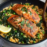 A skillet filled with orzo pasta, sautéed kale, and peas topped with two pieces of seasoned grilled salmon garnished with herbs and lemon wedges.