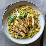 A bowl of fettuccine pasta topped with sliced grilled chicken, basil, and a pesto sauce, garnished with grated cheese.