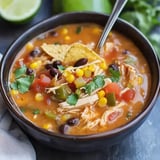 A bowl of chicken soup with black beans, corn, tomatoes, and cilantro, garnished with tortilla chips, accompanied by lime wedges in the background.