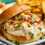 A close-up of a sandwich filled with shredded chicken, topped with cheese, bacon, and green onions, served with a side of French fries.