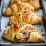 A close-up of golden-brown croissants filled with ham and cheese, garnished with chopped parsley on a baking tray.