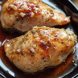 Two glazed chicken breasts topped with garlic and herbs are served on a black plate.