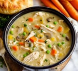 A bowl of creamy chicken soup with vegetables, including carrots, peas, and mushrooms, alongside a biscuit.