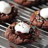 A close-up of chocolate cookies topped with melted chocolate, a dollop of whipped cream, and colorful sprinkles on a cooling rack.