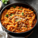 A close-up view of a pot filled with creamy fusilli pasta mixed with ground meat, topped with parsley and grated cheese.